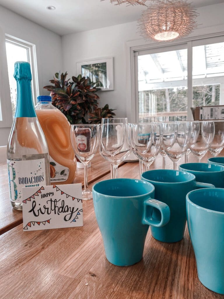 Wine glasses and mugs arranged on a wooden table along with a birthday card, bottle of wine and orange juice at this luxury staycation.