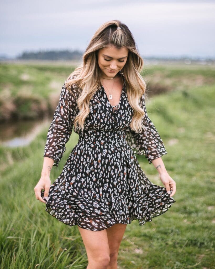 A girl wearing a black spotted dress. She has long blonde wavy hair. The grass is very green in the back ground and the sky is a grey blue colour.