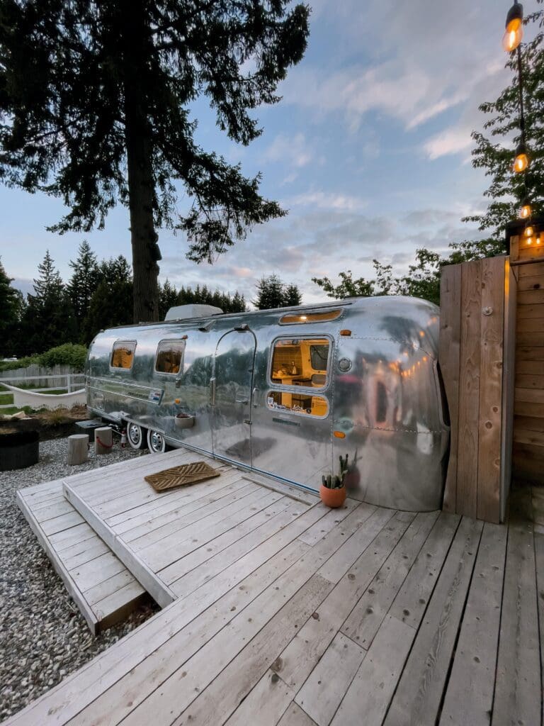 The view of the airstream at Tin Can Ranch from the deck that connects the outdoor shower, bath tub and sauna. off to the left is the fire pit and horseshoe game. The airstream is super shiny and the inside lights are glowing.