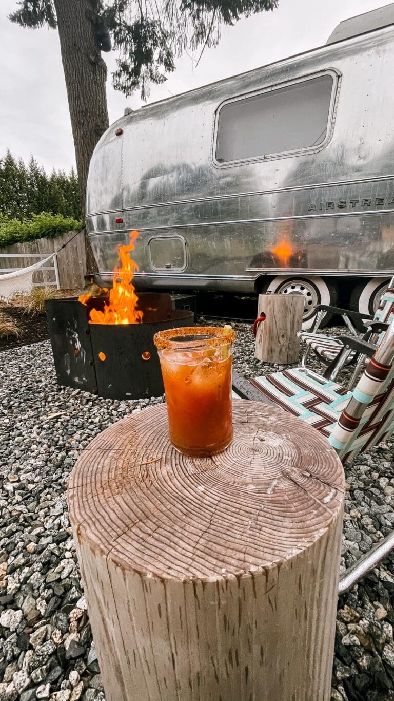 View of the fire pit next to the airstream at Tin Can Ranch with caesar front and centre on the log side table. The fire is burning high and is reflecting in the shiny airstream