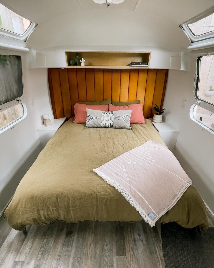 A view of the bed at Tin Can Ranch inside of the airstream. The bedding is a light frost green colour and the pillows are a pink salmon colour. The head board is a dark tan colour.