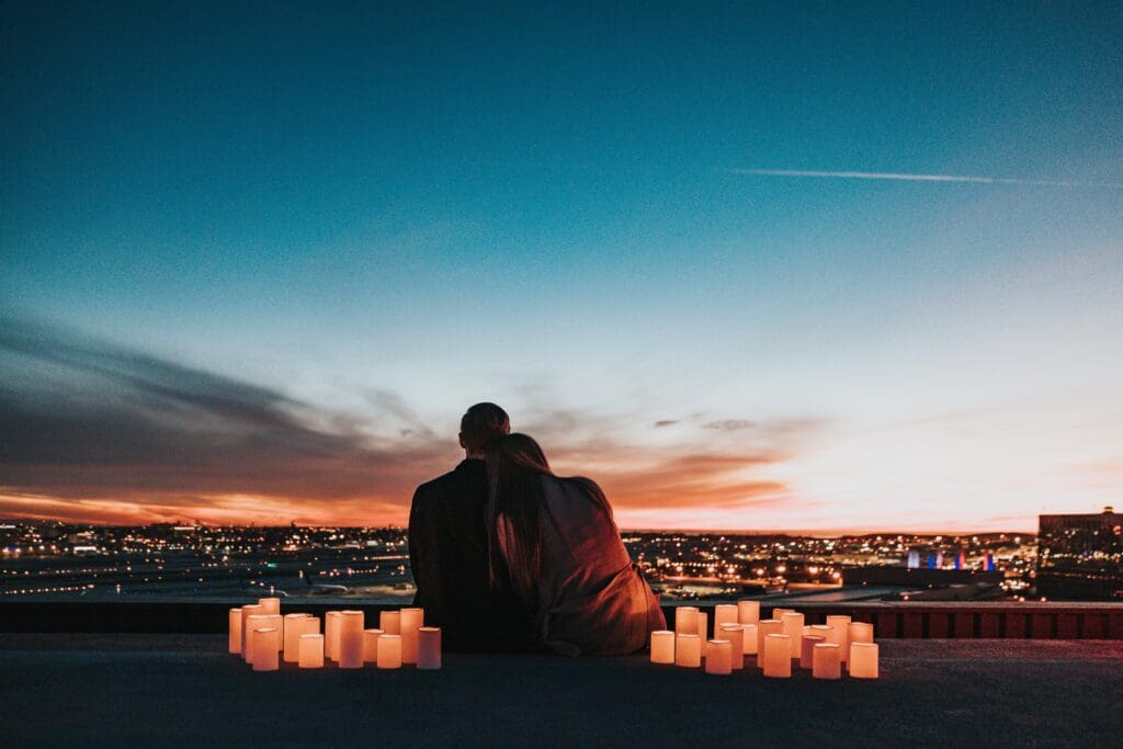 A couple sitting on a high cliff overlooking the city. They have candles on either side of them. They are a dark silhouette
