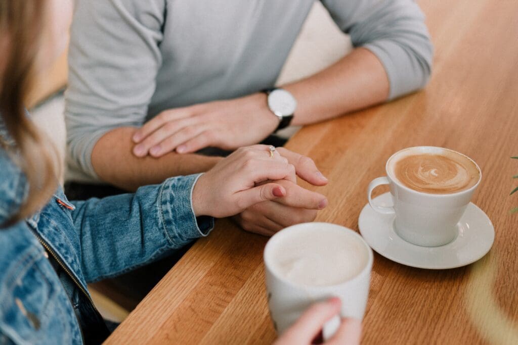 A couple holding hands at a table with coffee. The man is wearing a watch and grey shirt. The lady is wearing a jean jacket.and holding the coffee mug.