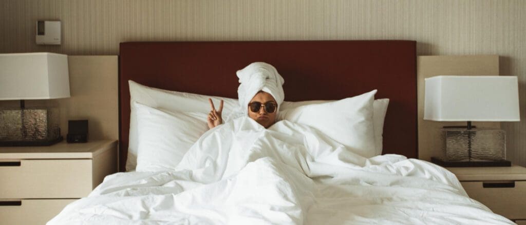A girl curled up in bed in a hotel room. She has a towel on her head and throwing up the peace sign. She has brown sunglasses on. The sun is coming in through the side window.
