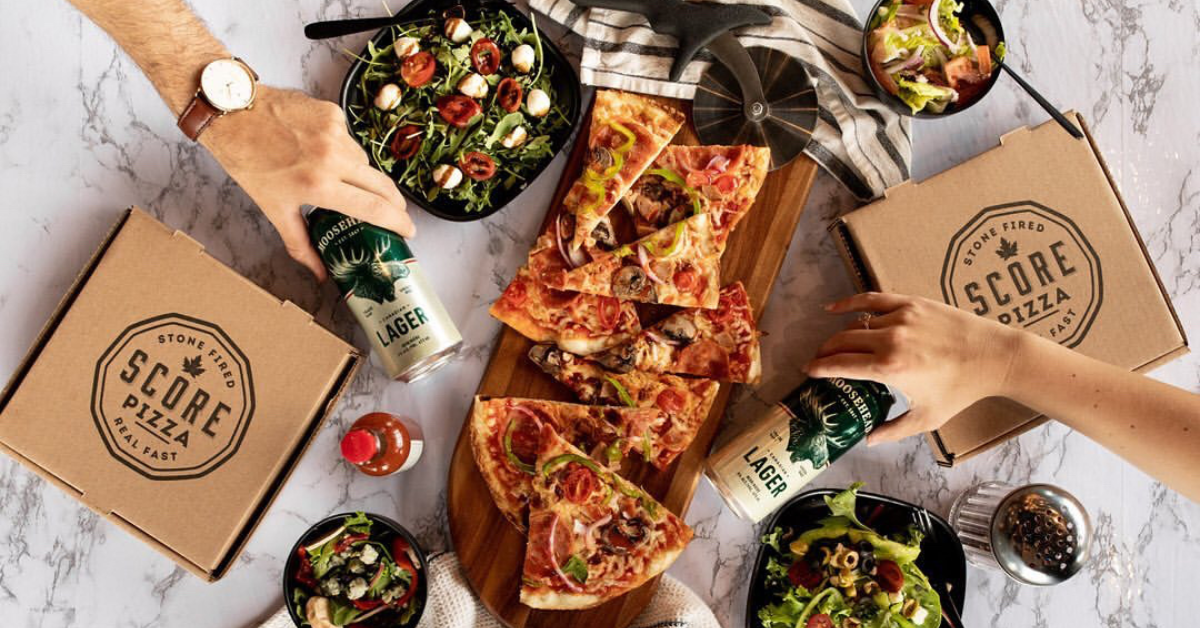 Overhead photograph of a spread of Score Pizza slices, salads, and hands holding cans of beer.