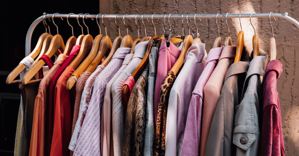 shirts, sweaters, and jackets hanging on a white clothing rack behind a brown/tan cement background