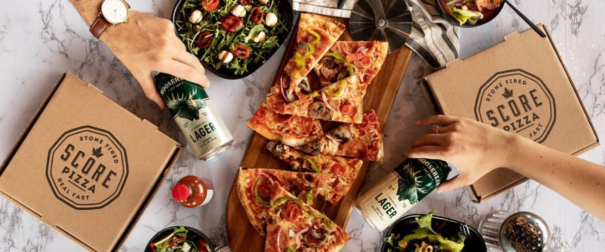 Overhead photograph of a spread of Score Pizza slices, salads, and hands holding cans of beer.
