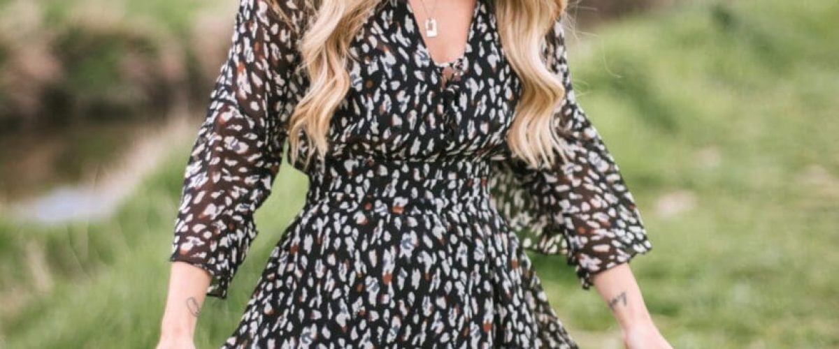 A girl wearing a black spotted dress. She has long blonde wavy hair. The grass is very green in the back ground and the sky is a grey blue colour.