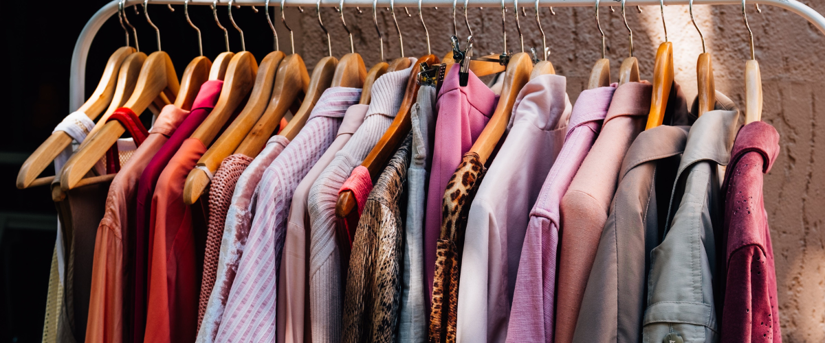 shirts, sweaters, and jackets hanging on a white clothing rack behind a brown/tan cement background