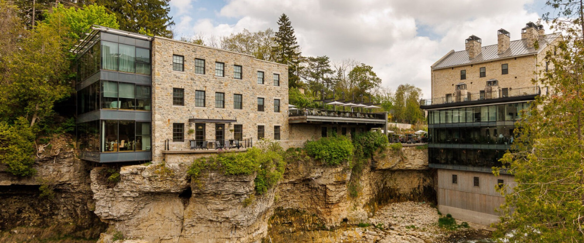 Elora brick cottage on the side of a cliff with a blue river flowing underneath and green cedar trees in the background