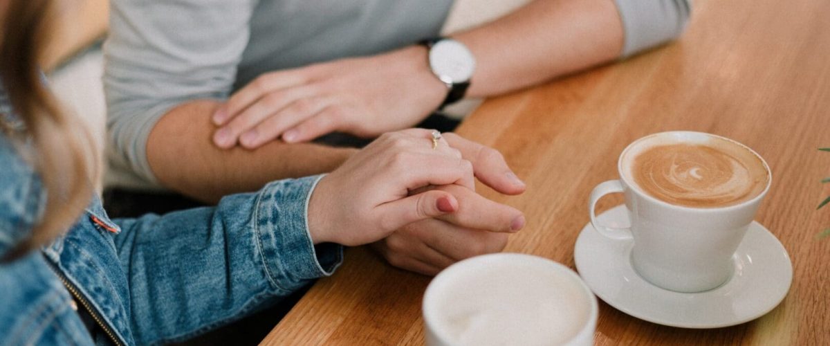 A couple holding hands at a table with coffee. The man is wearing a watch and grey shirt. The lady is wearing a jean jacket.and holding the coffee mug.
