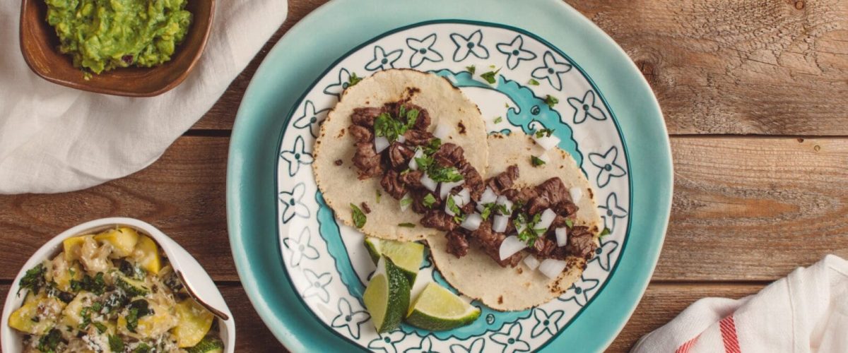 An arial view of a taco dinner spread. The table is medium to dark wood. There are three tacos in the centre. guacamole is off to the left in a bowl and a pineapple salsa in the bottom left.